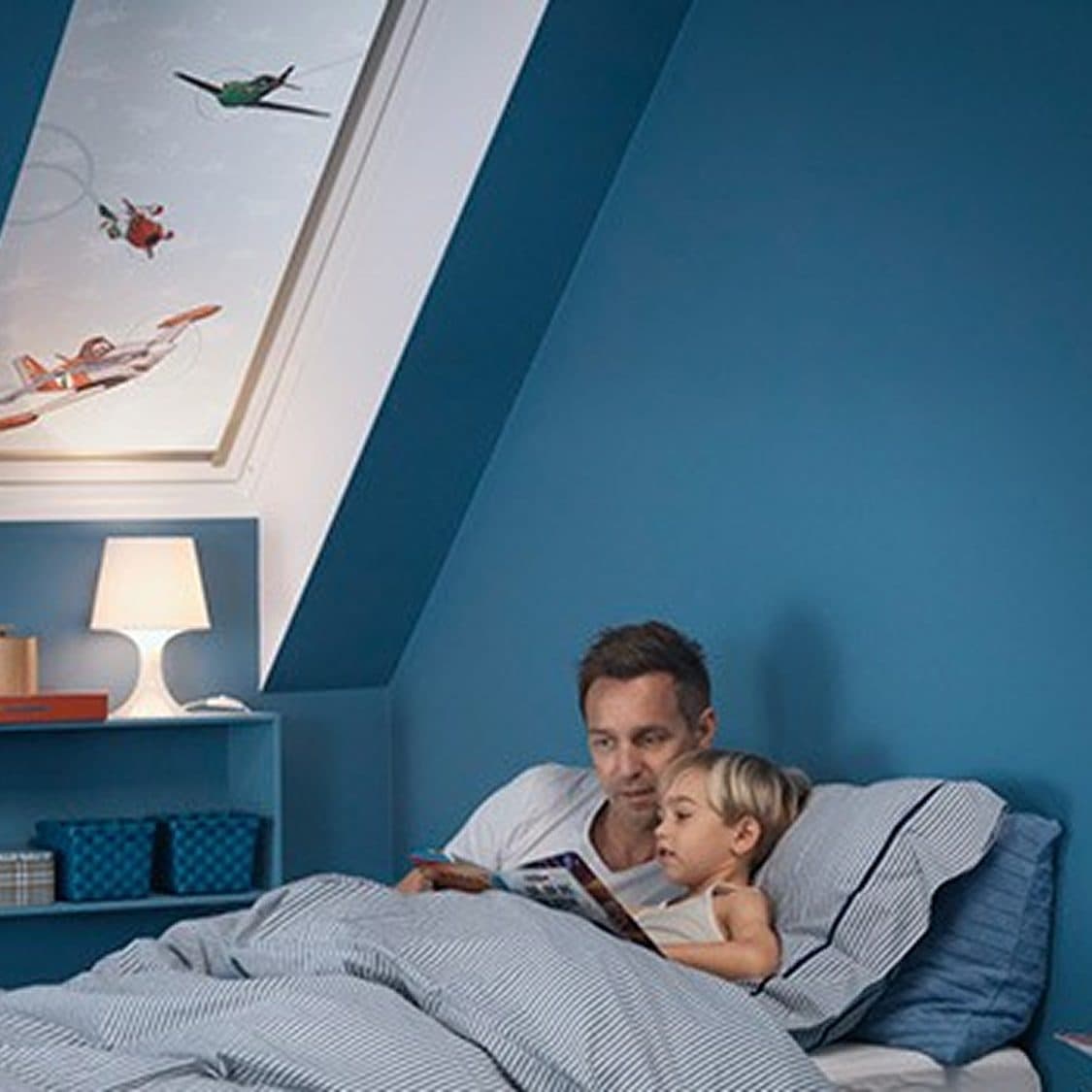 An adult reading to a small child in bed in a blue attic bedroom, a roof window above them and a lamp lit on the shelf.