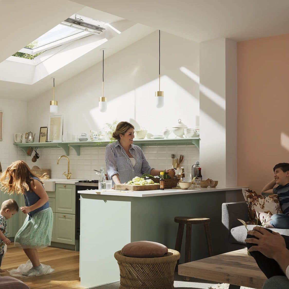 A woman preparing food at a kitchen island under a roof window while two children play nearby in the morning light.